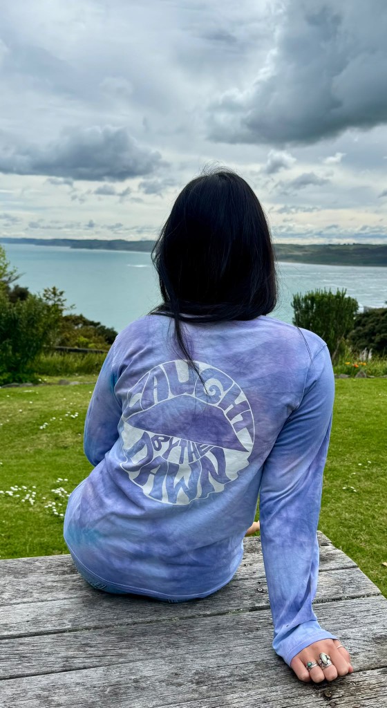 Back view of the Caught by the Dawn tie-dye long-sleeve tee with logo, worn outdoors on a coastal picnic table