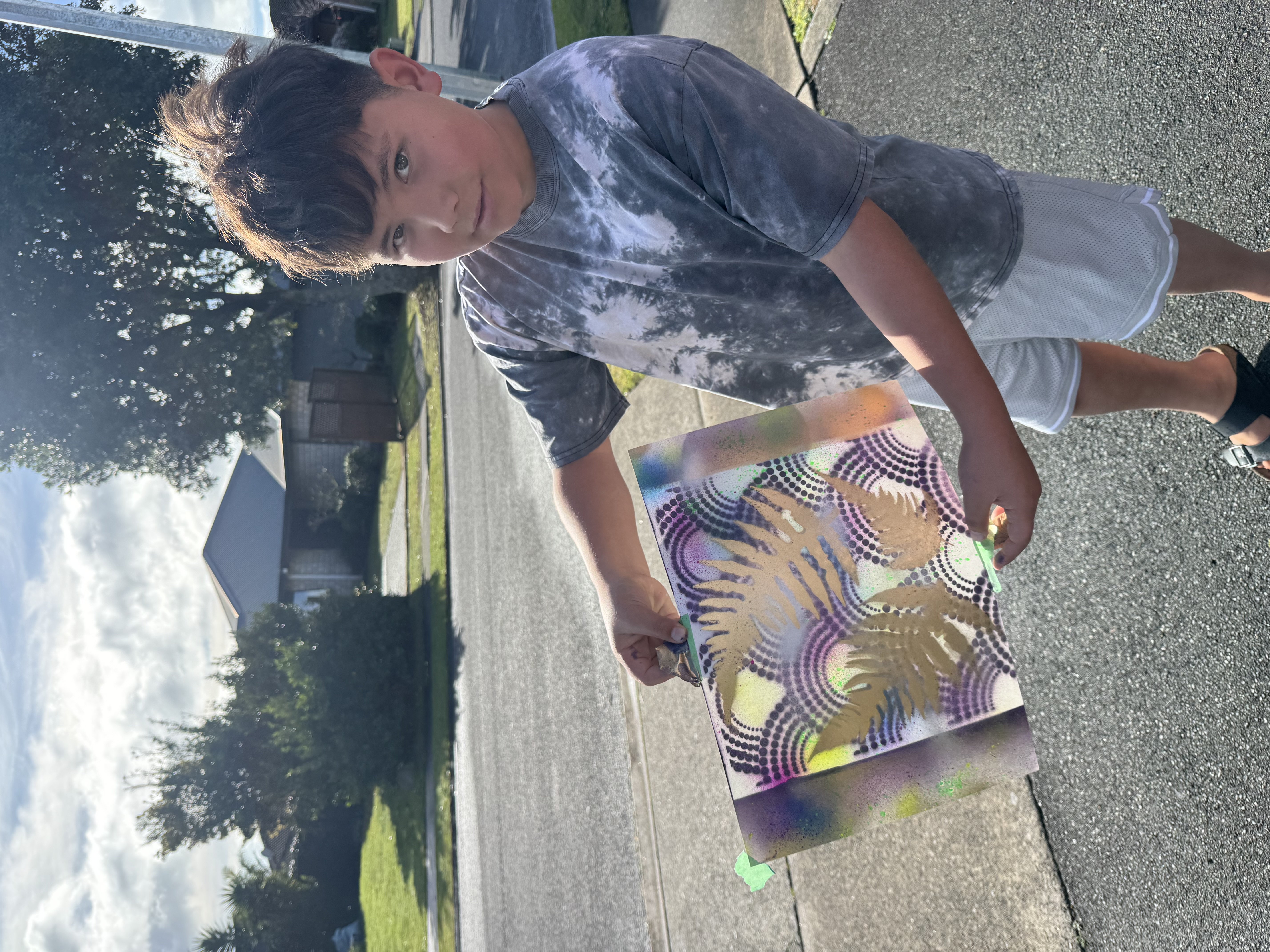 Boy holding up spray-painted fern leaf artwork outdoors