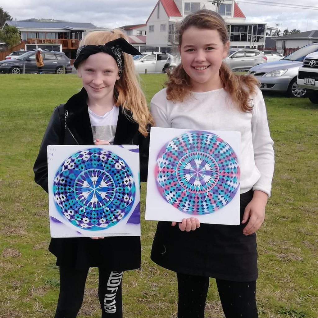 Two young girls displaying geometric mandala paintings on canvas in a field