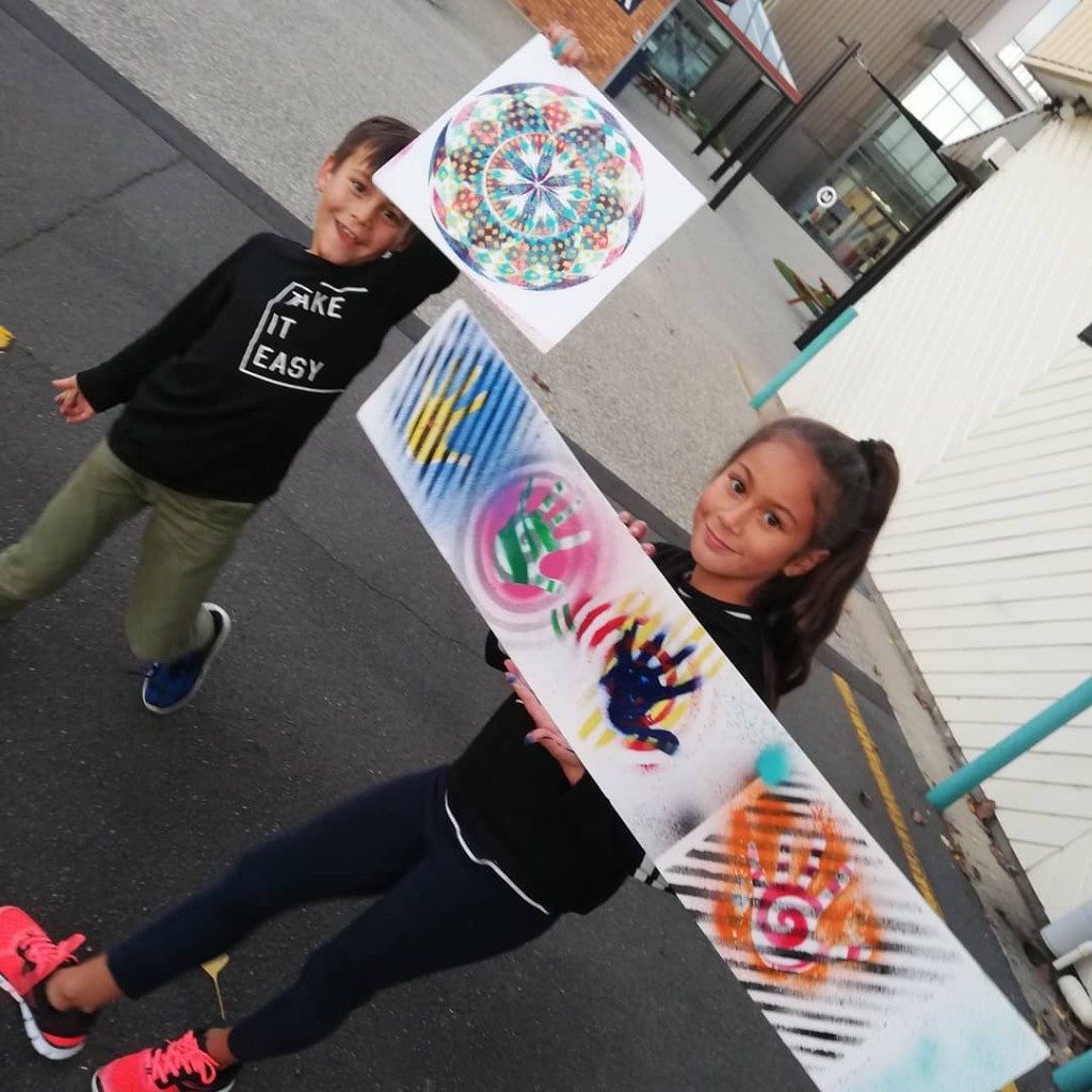 Two children smiling and holding colorful mandala and handprint artwork outdoors