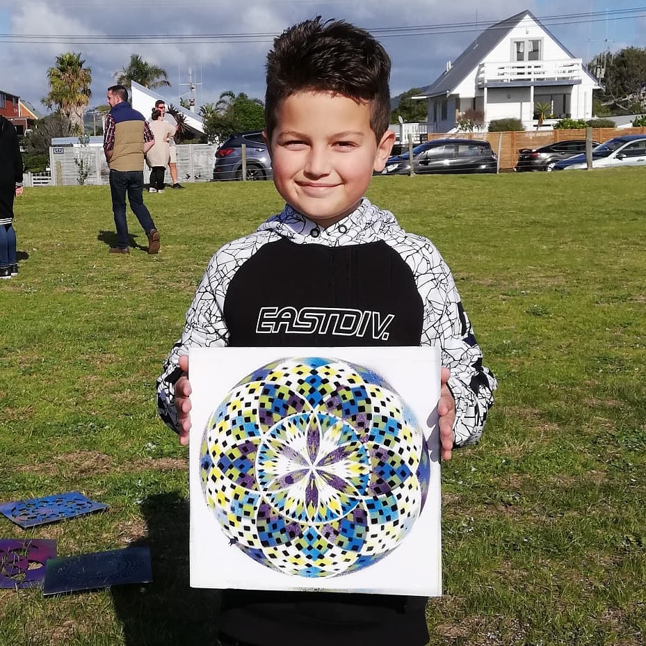 Boy proudly holding a colorful geometric mandala painting on canvas in a sunny field