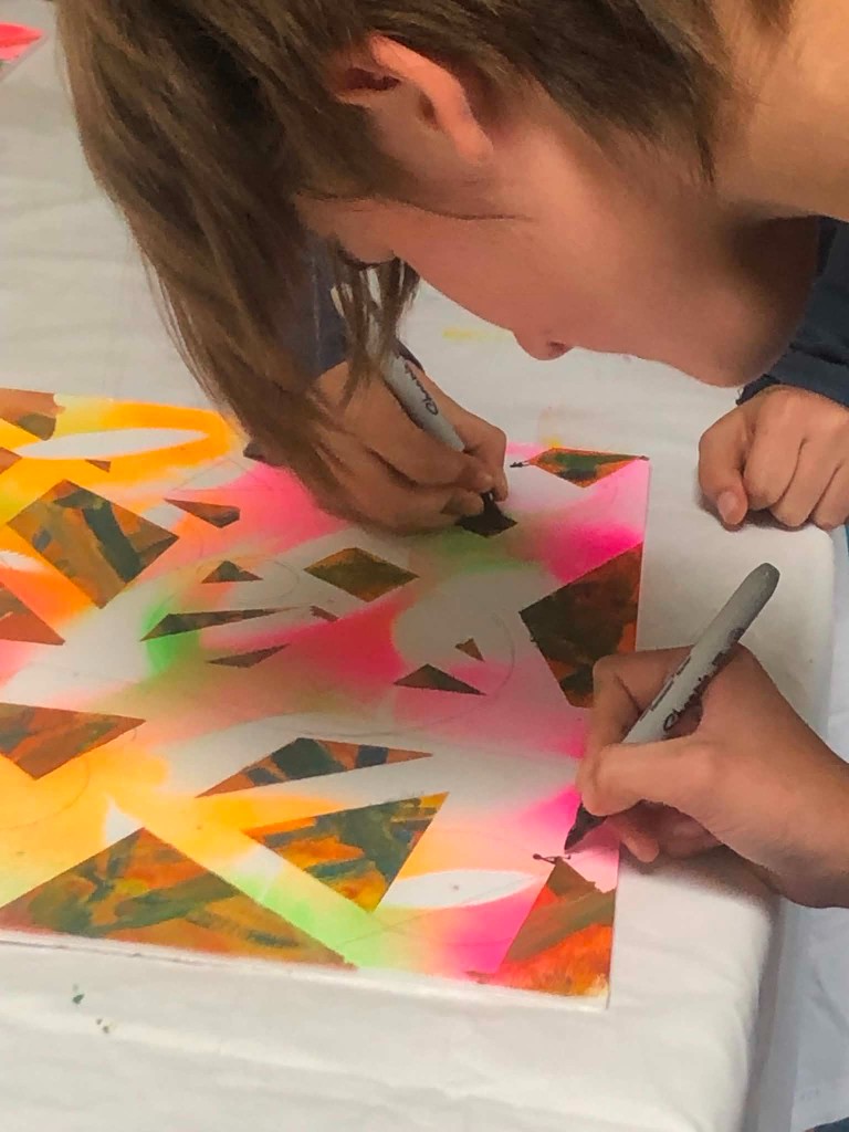 Hands drawing detailed lines on a bright geometric artwork with markers