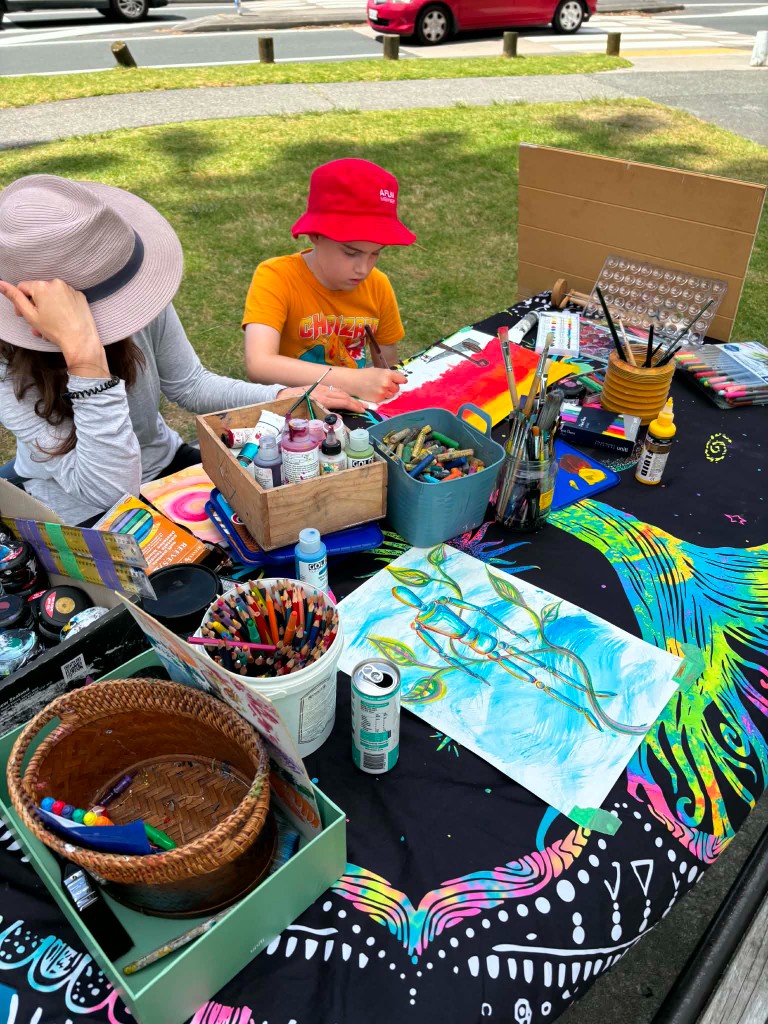 Facilitator and child painting together at an outdoor table covered in art supplies