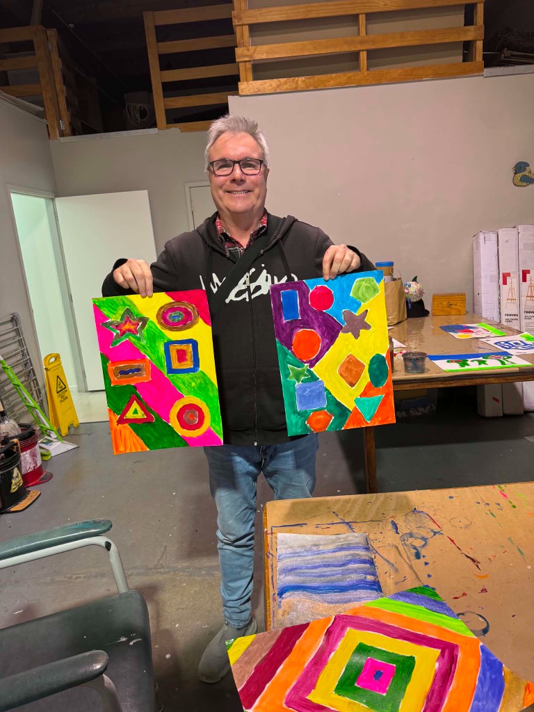 Artist holding two colorful geometric paintings in an art studio
