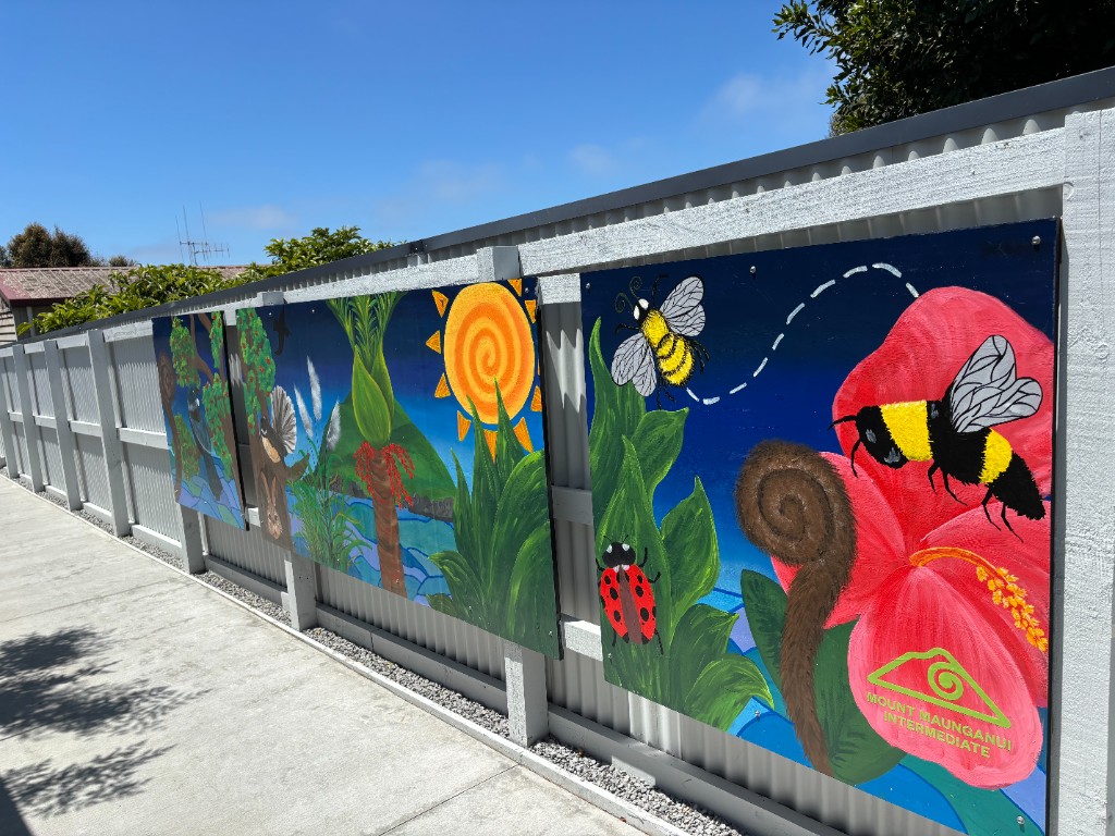 Student mural panels mounted on a fence featuring native flowers, bees, and Mount Maunganui Intermediate branding